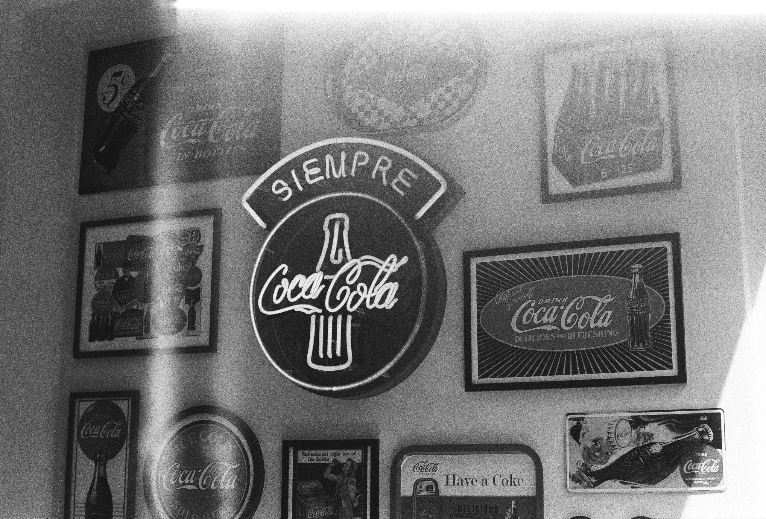 Black and white photo of a wall display with vintage Coca-Cola signs, including the iconic Coca-Cola bottle and logo designs
